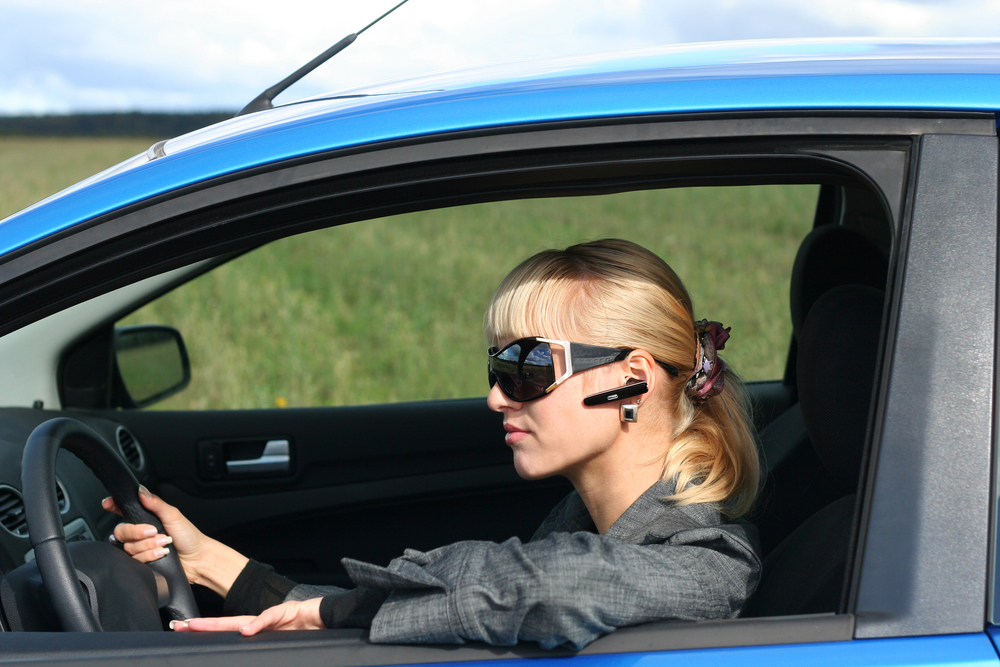 young blond woman in a blue car in sun-glasses with hands free bluetooth