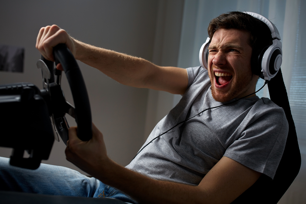 technology, gaming, entertainment and people concept - young man in headphones with pc computer playing car racing video game at home and steering wheel