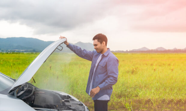 A young man looking into the hood of his car as smokes comes out.