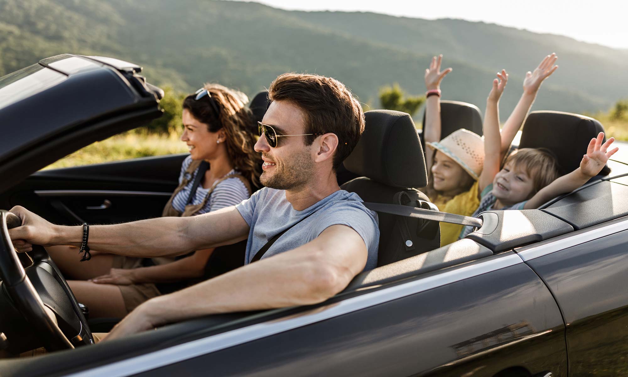 A family of four driving down the road in a convertible on a sunny day.