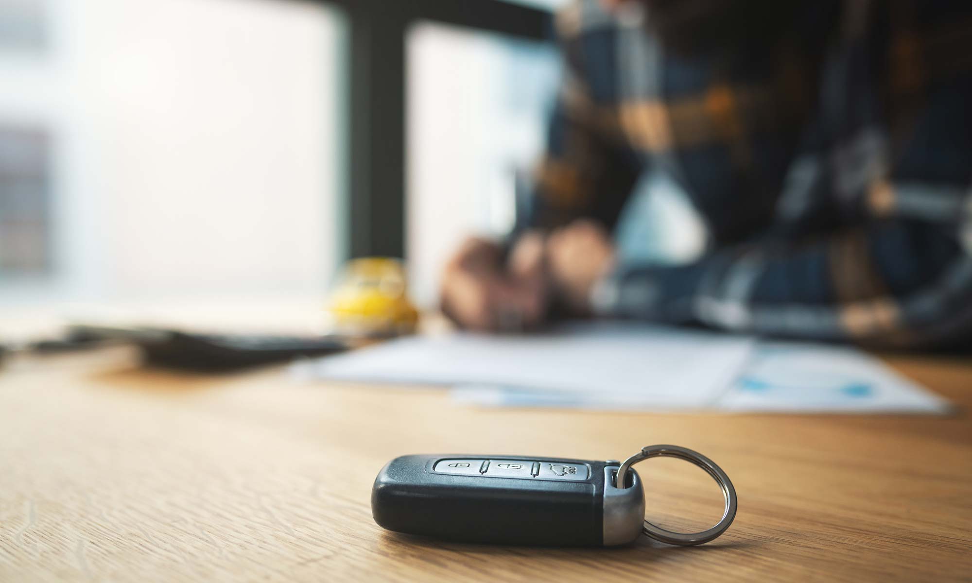 A car key fob sits on a table as a man signs a car warranty contract in the background.