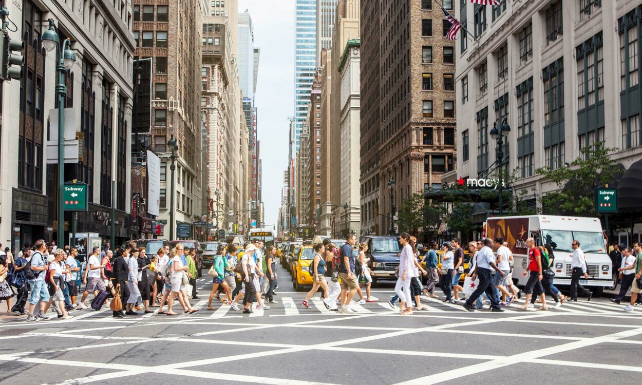 A large group of pedestrians crossing in front of a row of cars at a New York City crosswalk.