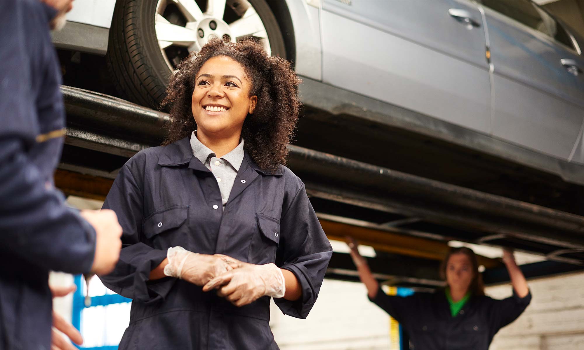 A female mechanic speaks with a customer while her colleague work on a vehicle behind her.s