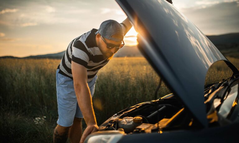 A young man in shorts and a t-shirt leans over the open hood of his car.