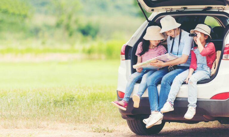 A mother and her two young daughters, all wearing hats, are sitting in the back of their open car trunk looking at a road trip map.