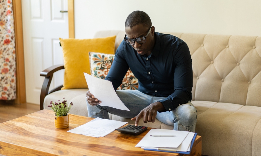 Man sitting on a sofa is comparing documents while using a calculator in his living room