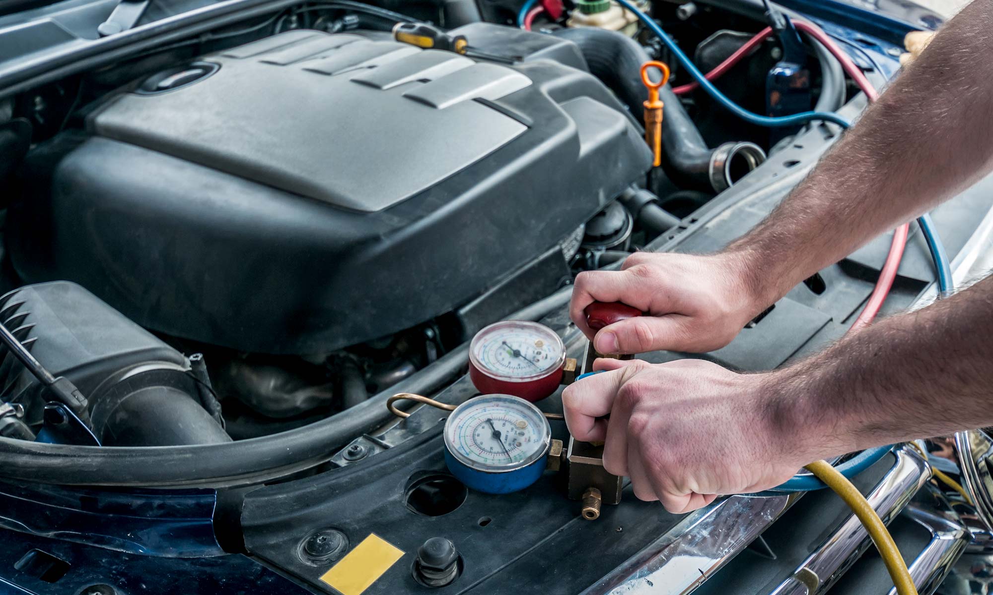 A male car mechanic's hands placing two Freon charging cables to a car's engine.