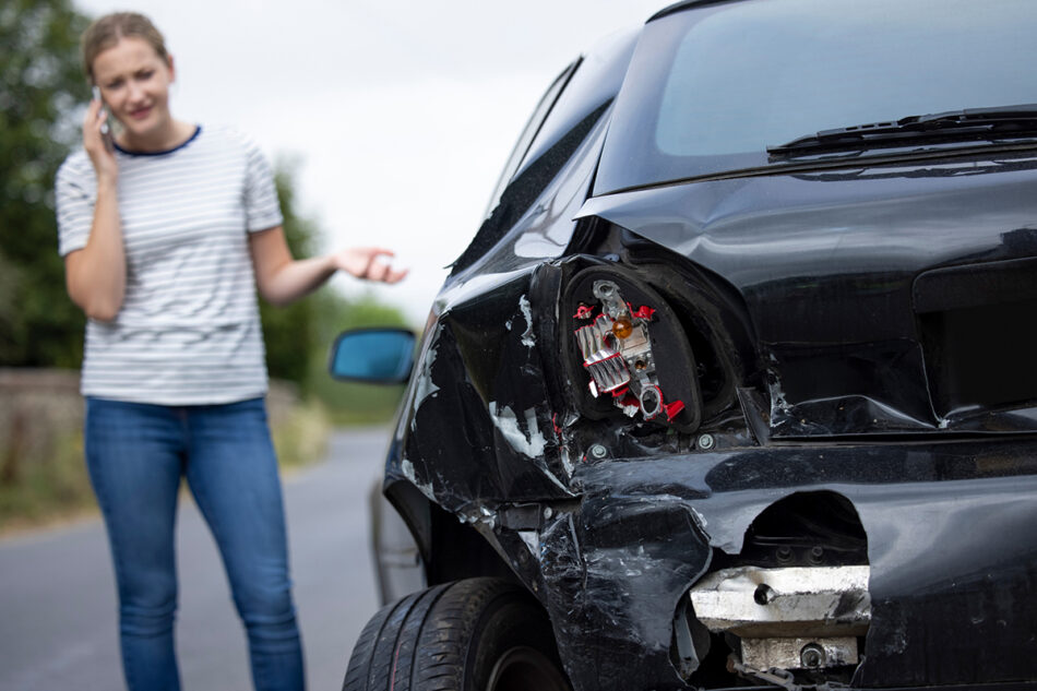 A woman standing in front of her damaged car talking on the phone.