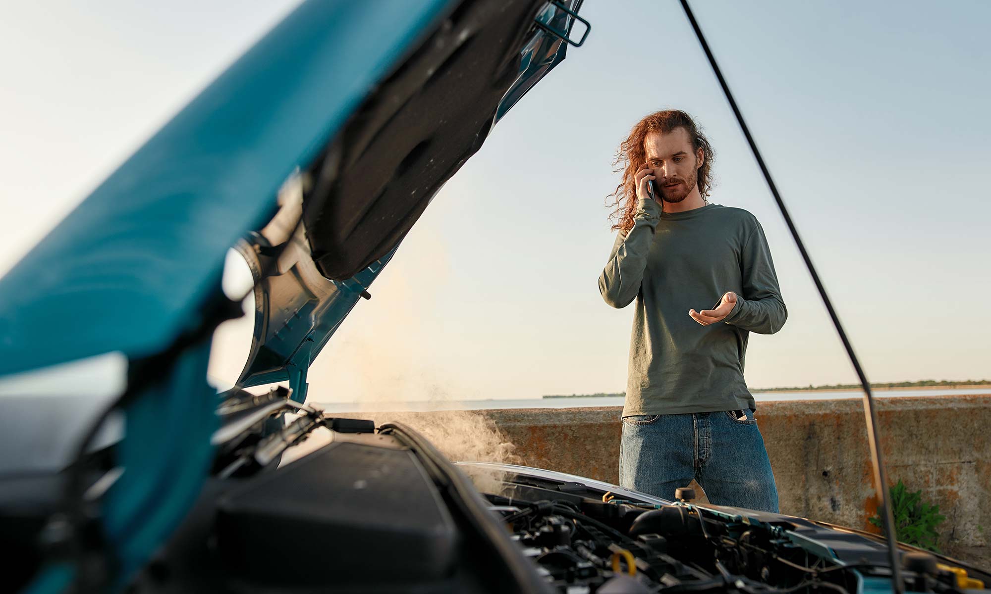 A man talking on the phone as he standing next to his open car hood with the engine smoking.