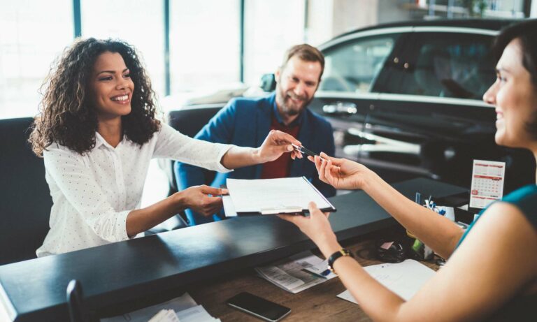 A couple handing over paper work to a salesman at a car dealership.