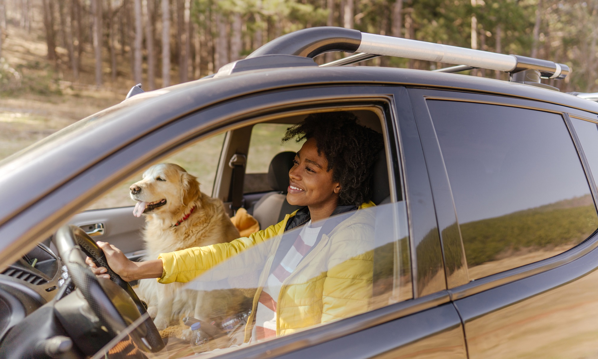 Photo of a young smiling woman driving a car. Her dog is sitting on a passenger seat