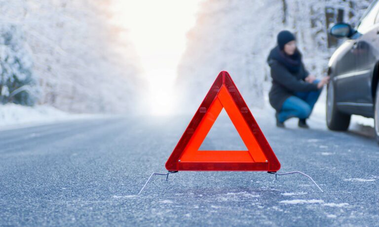 A woman kneeling next to her car in the winter with an emergency triangle reflector in the road.