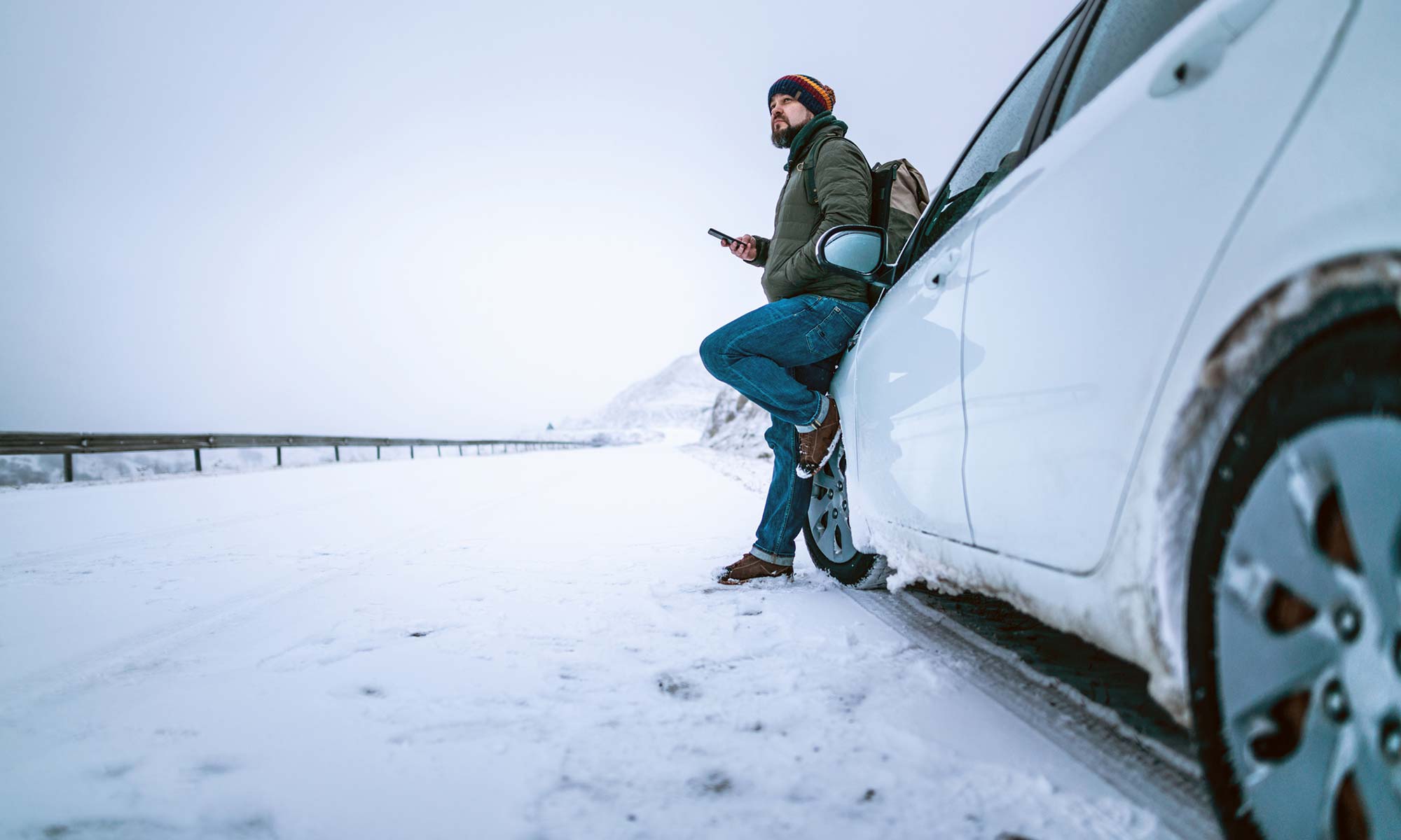 A young man leaning against his car on the side of a snowy road.