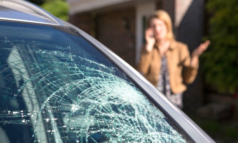 A woman talking on the phone next to her car's cracked windshield.