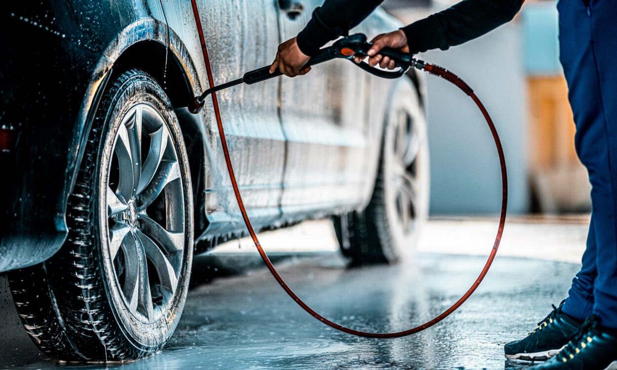 A man using a hose with a cleaning attachment to clean his car's tire.