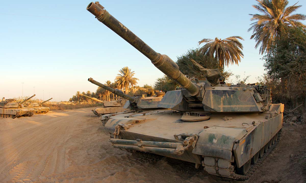 A row of M1 Abrams tanks parked in the sand.