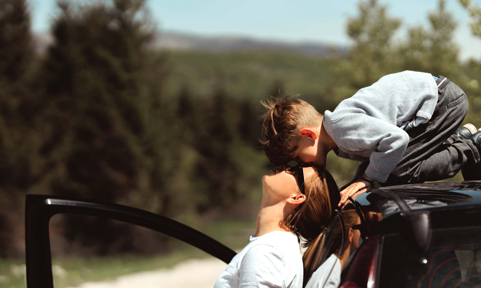 A young child on top of a car kissing his mother's forehead.