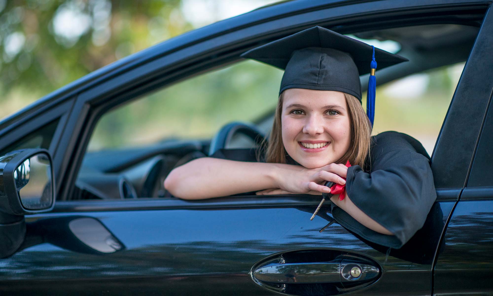 A young girl in a high school graduation cap & gown smiles in her new car.