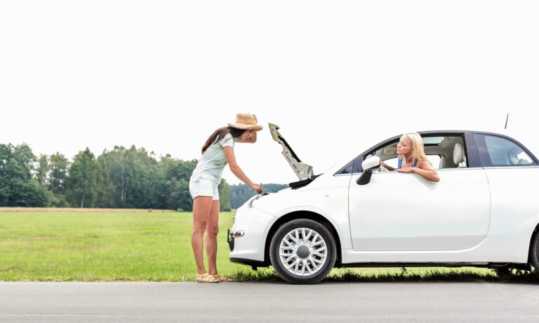 A young women checks under the hood of a car while her friend is looking out the window from the driver's seat.