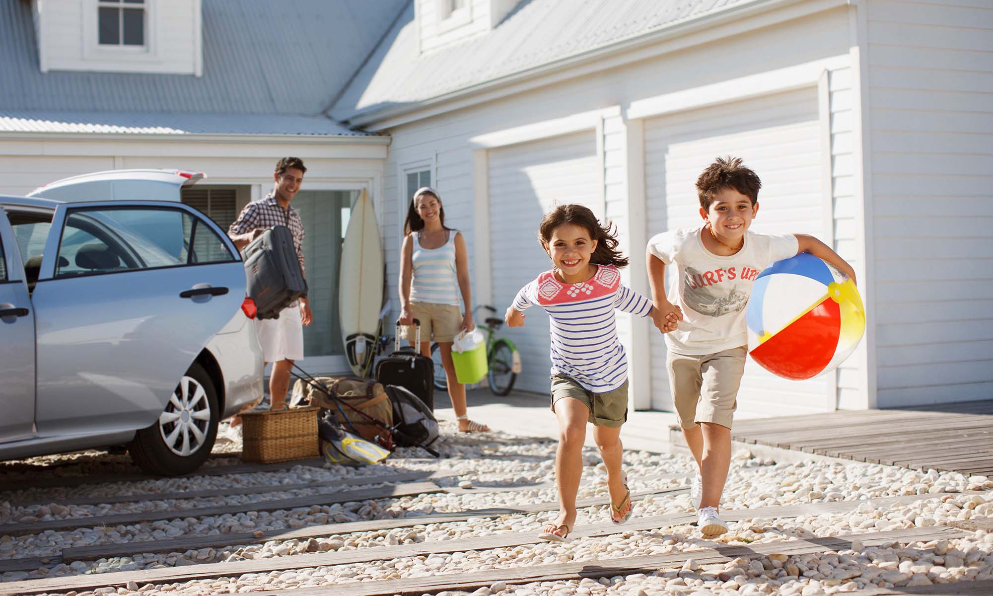 A young brother and sister run toward the camera while their parents unpack their vehicle at a vacation home.