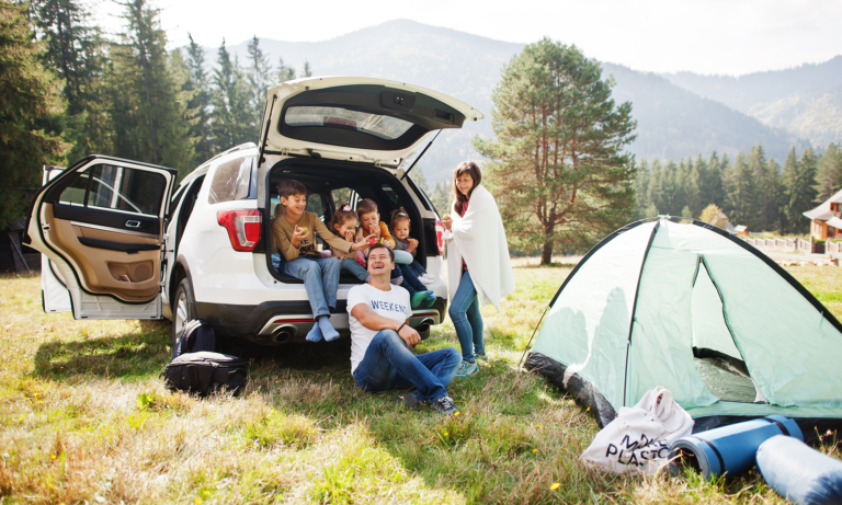 A family of 6 sitting in the open trunk of their car on a grassy field with a tent set up in front of them.