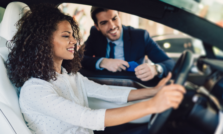 A young woman sitting in the front seat of a car while a salesman talks to her from the window.