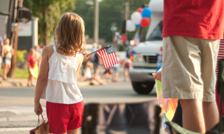 A young girl holding a small American flag next to her father as they watch a 4th of July parade.