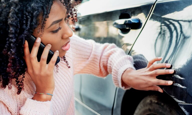 A young African American woman looks at scratches on her car while talking on her cell phone.