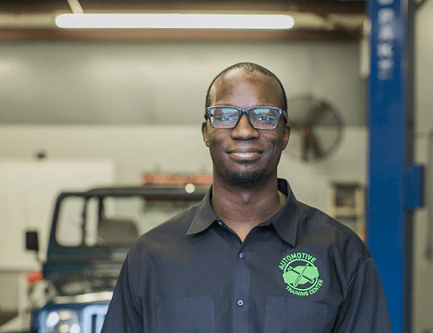 Certified auto technician Larry Witherspoon Jr. stands in an auto body shop with a car behind him