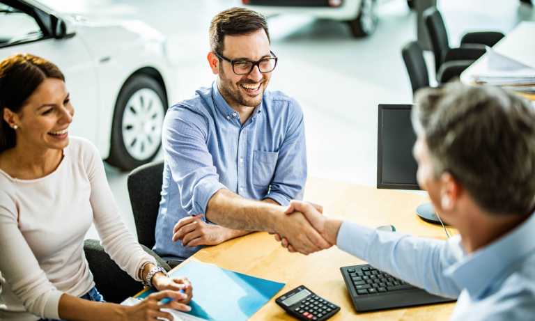 A young couple shaking hands with a car salesman after purchasing a new car.