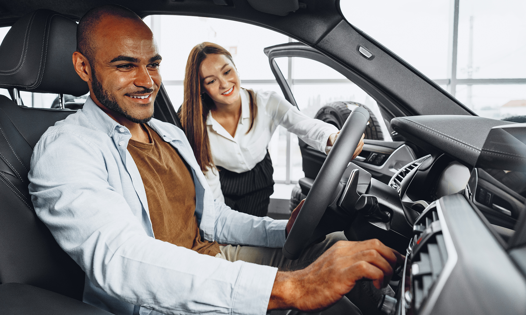 A young man sitting in the front seat of a car that he is test driving, while a young saleswoman shows him features on the car's dashboard.