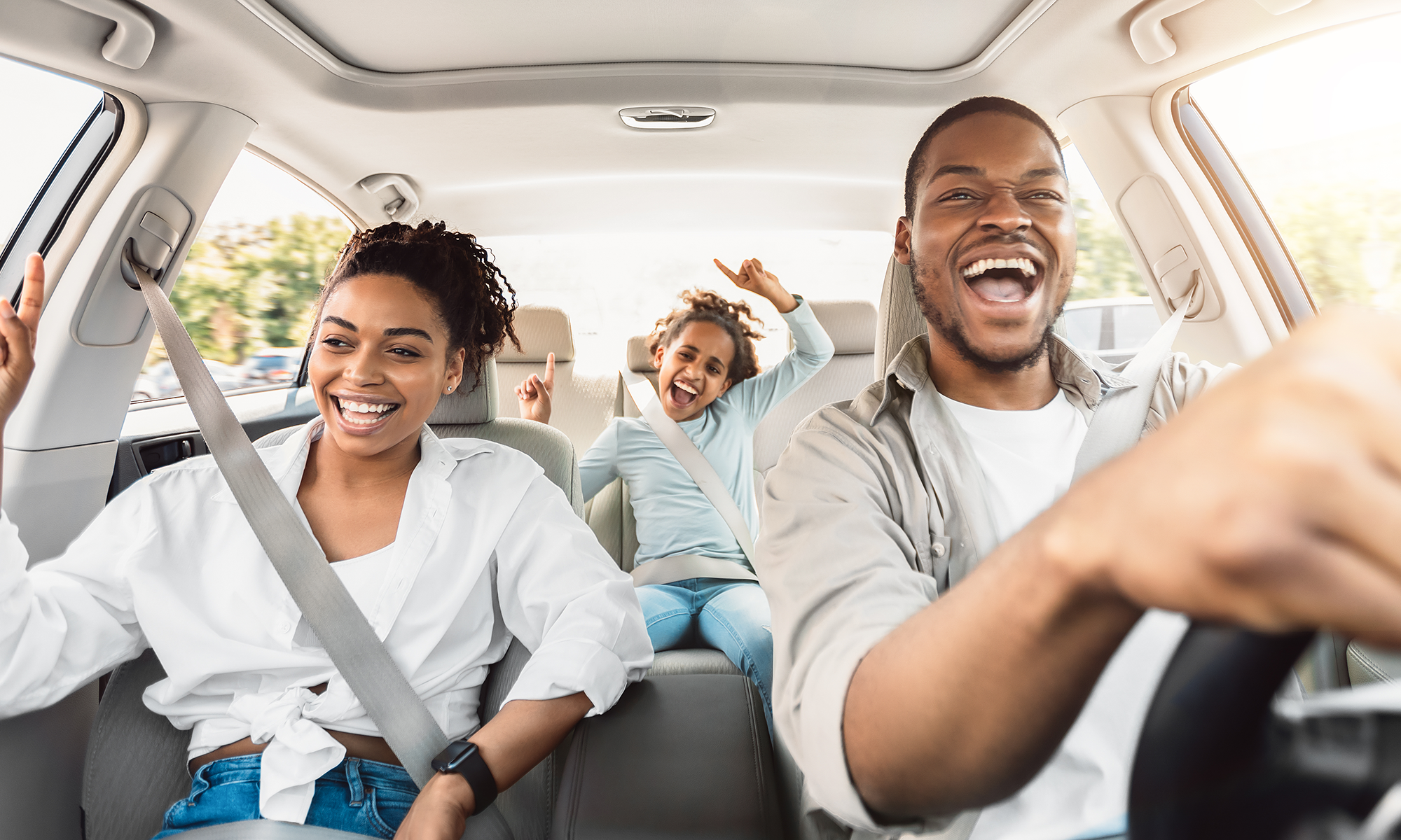 A young African American couple and their daughter laughing and singing while driving in their car.