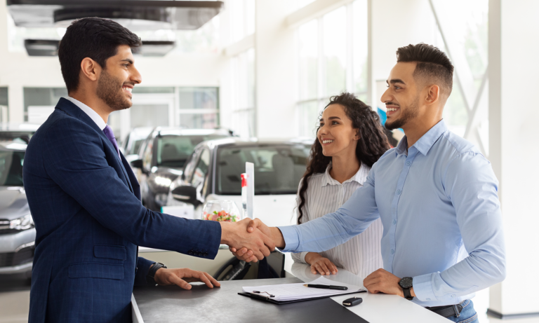 A young couple shaking the hand of a car salesman after trading in their old vehicle.