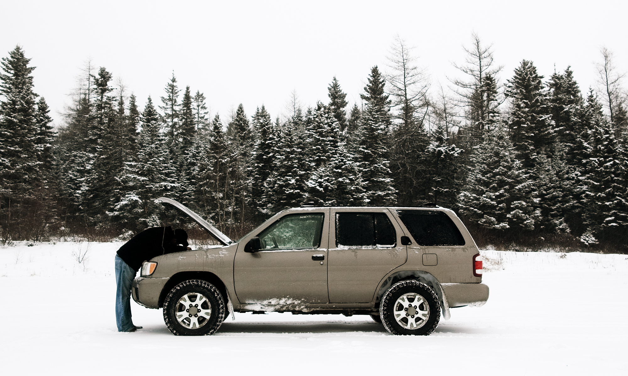 A car owner looking under the hood of their tan SUV in the middle of a snowy field with trees behind them.