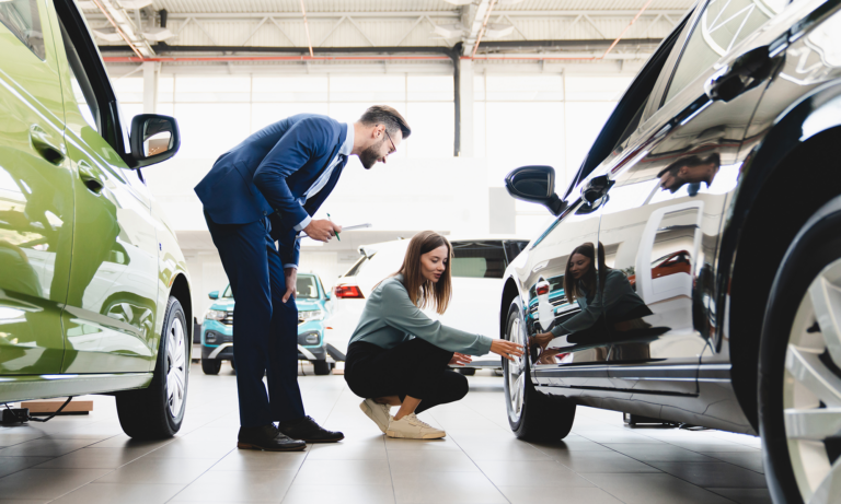 A young woman kneeling down and inspecting the front driver-side wheel of a car she is considering purchasing while a salesman stands next to her.