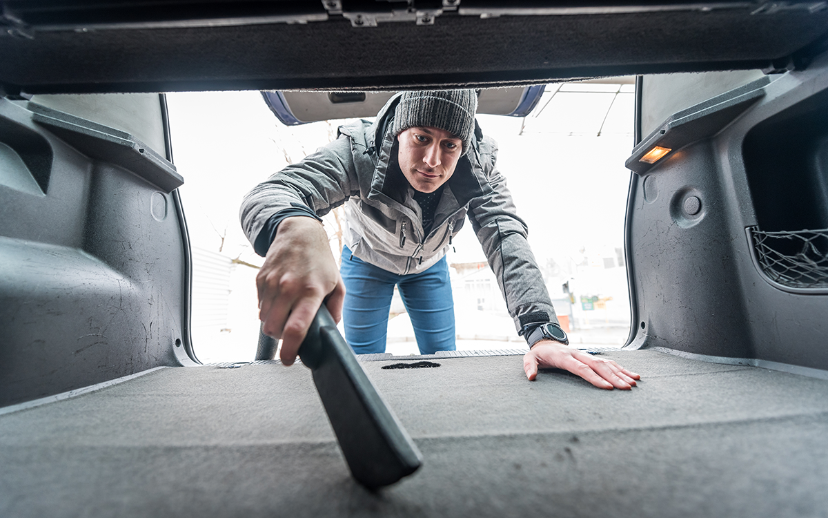 A man dressed in a winter coat and hat vacuums out the trunk of his car. 