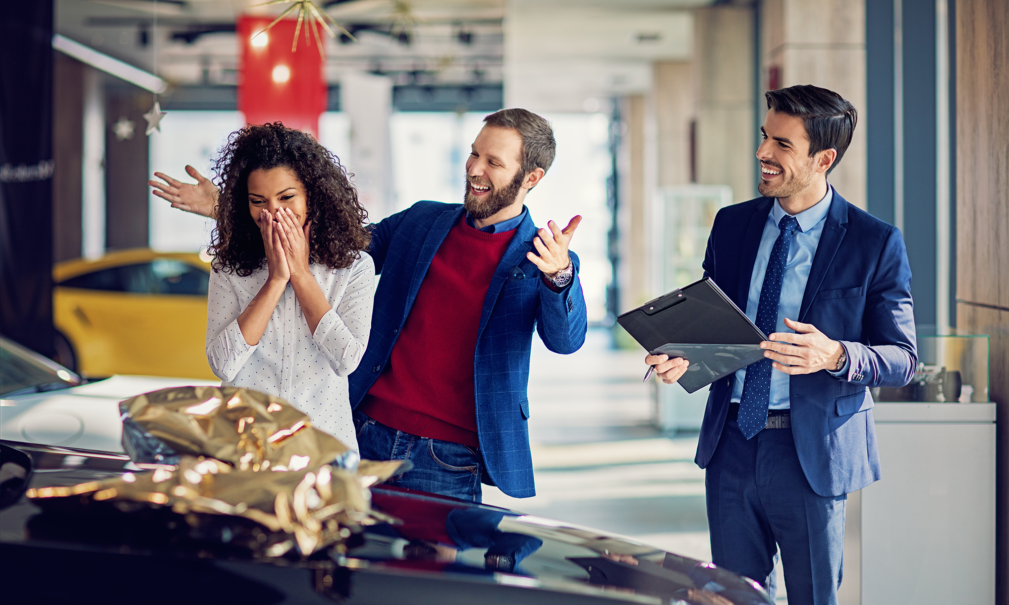 A husband surprises his wife with a new car that has a bow on top while a car salesman stands nearby.