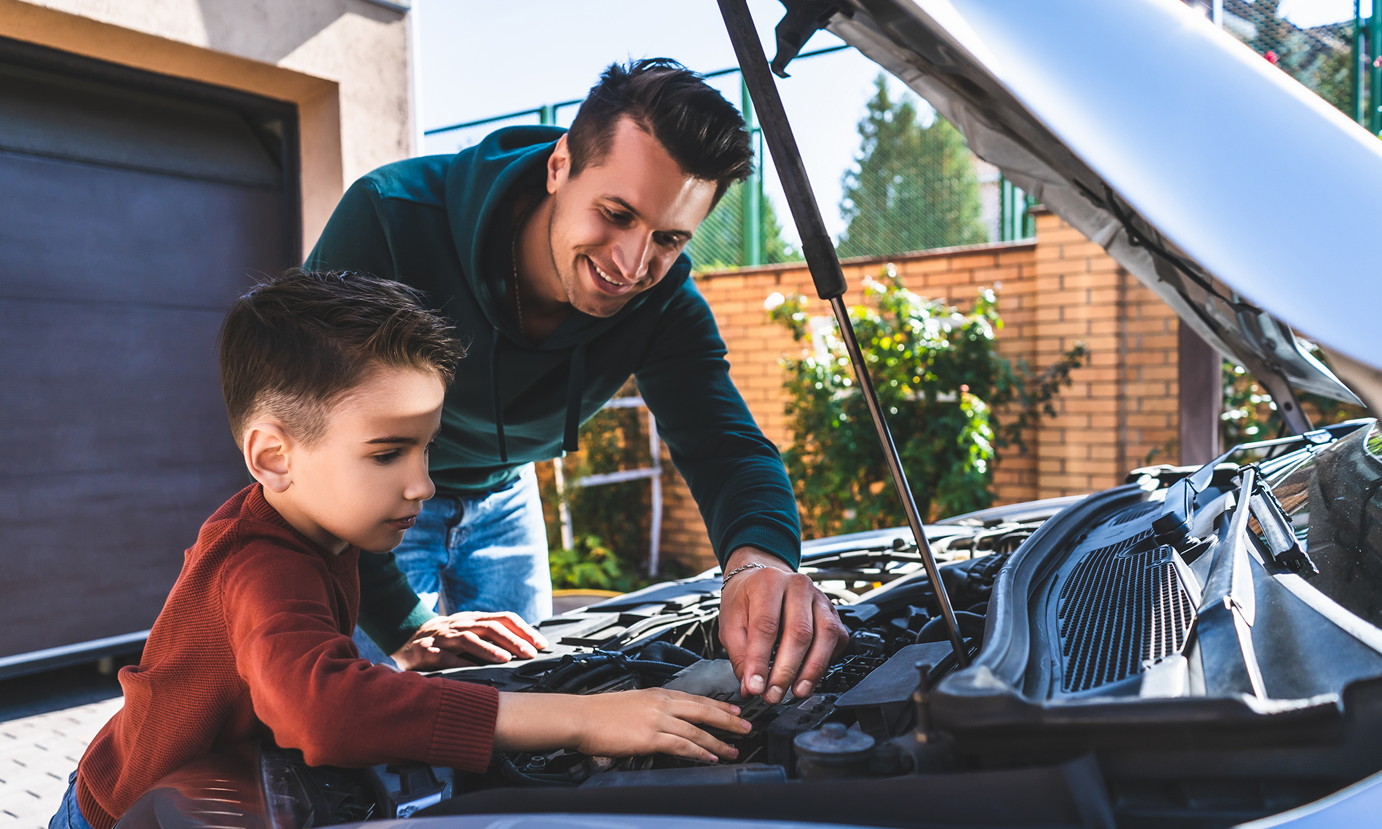 A father and young son work on their car in their driveway.