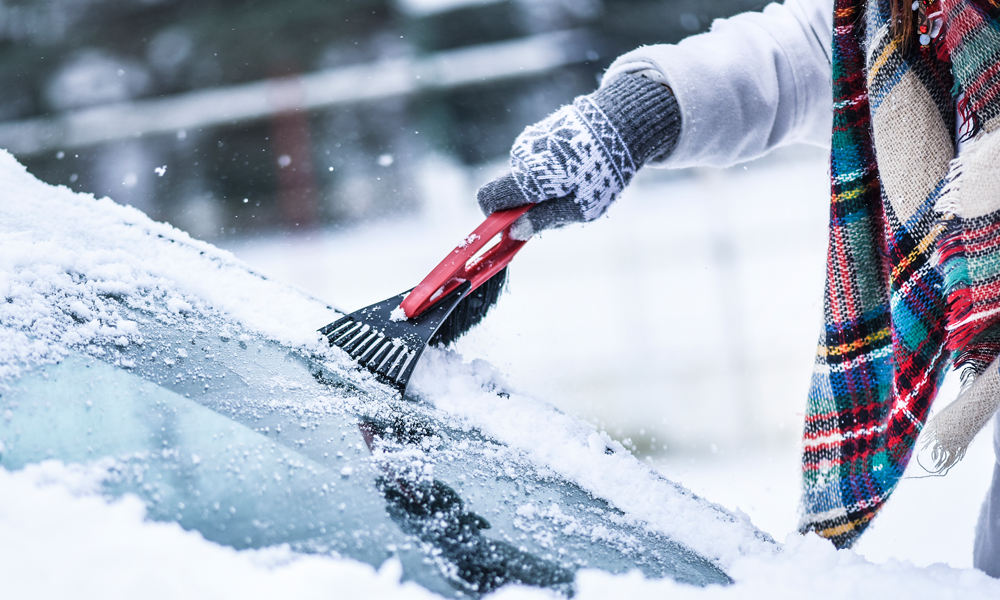 A close-up of a person scrapping their car's windshield with an ice scrapper while wearing clothes, a scarf and a white winter jacket.