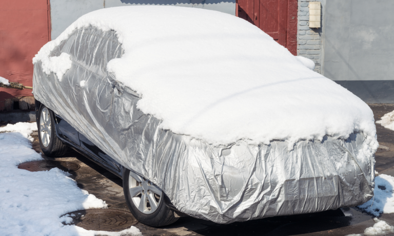 A car parked outside with a silver car cover and a layer of snow on top of it. A car cover can be a great thing to use when storing a vehicle during the winter when you do not have access to a garage.