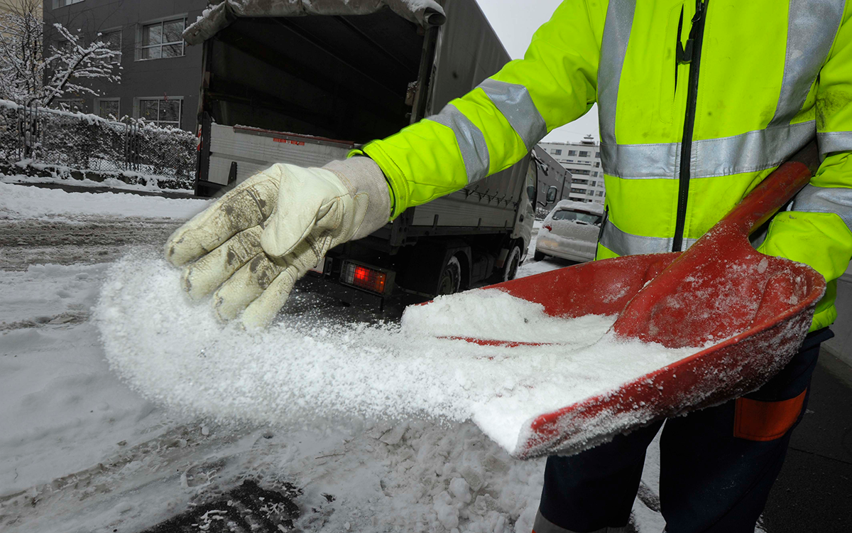 A close-up of a worker spreading road salt with a shovel on a snowy road.