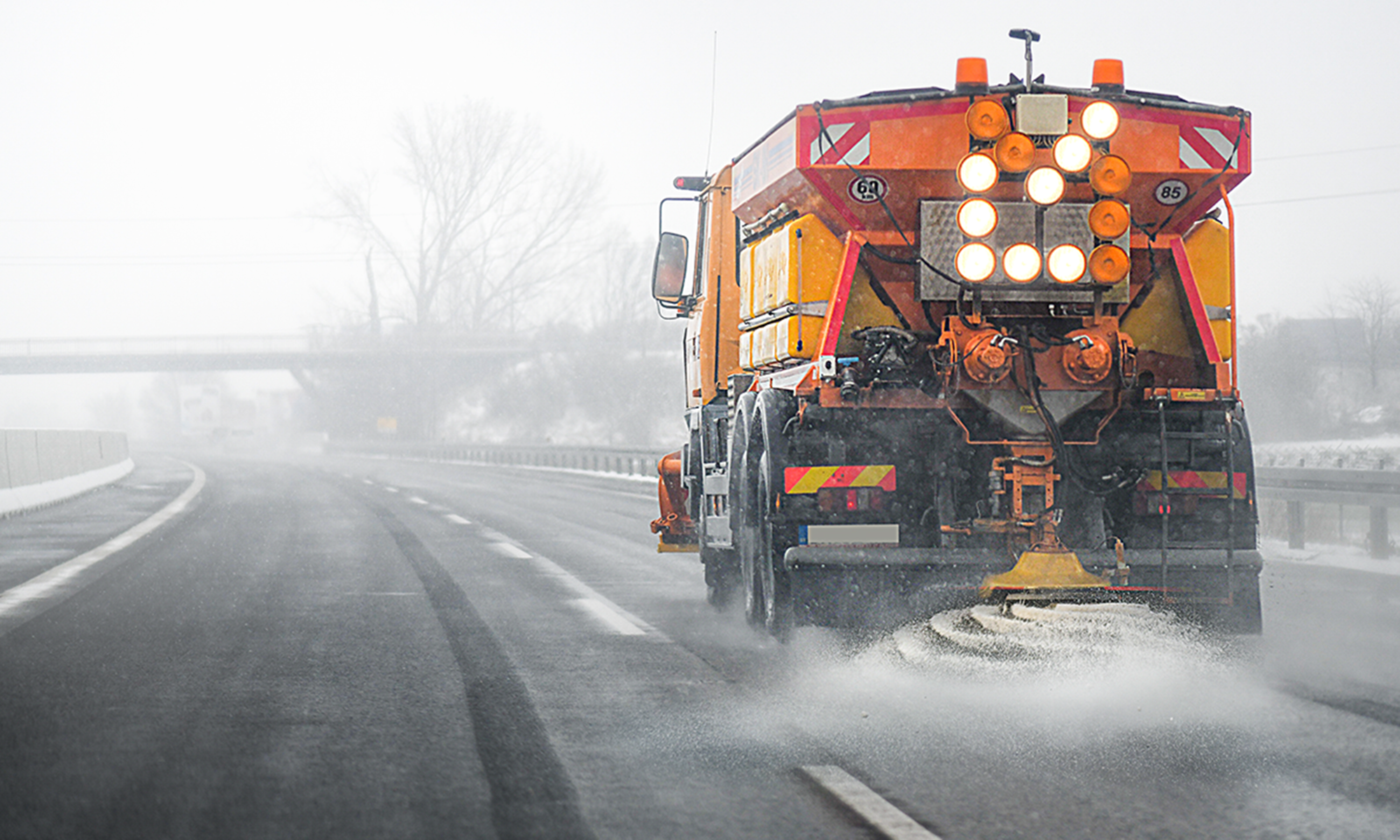 A snow blow driving down a rural highway spraying road salt out of the back.