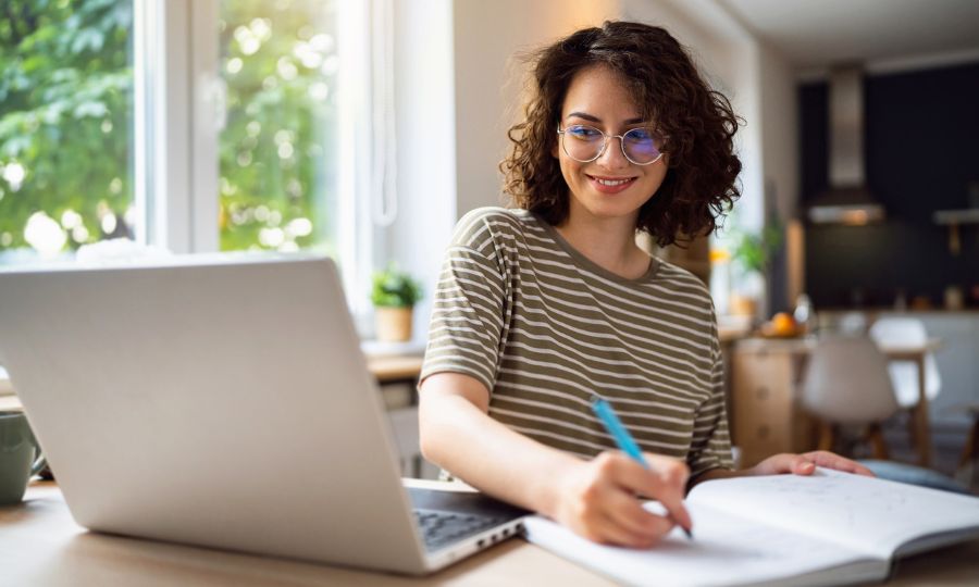 Young woman sitting at her laptop and writing in a notebook